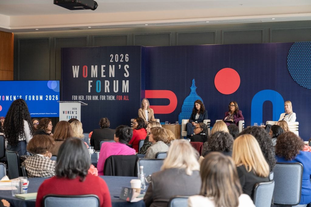 Four women sit on the dais during a panel discussion in front of a full audience.