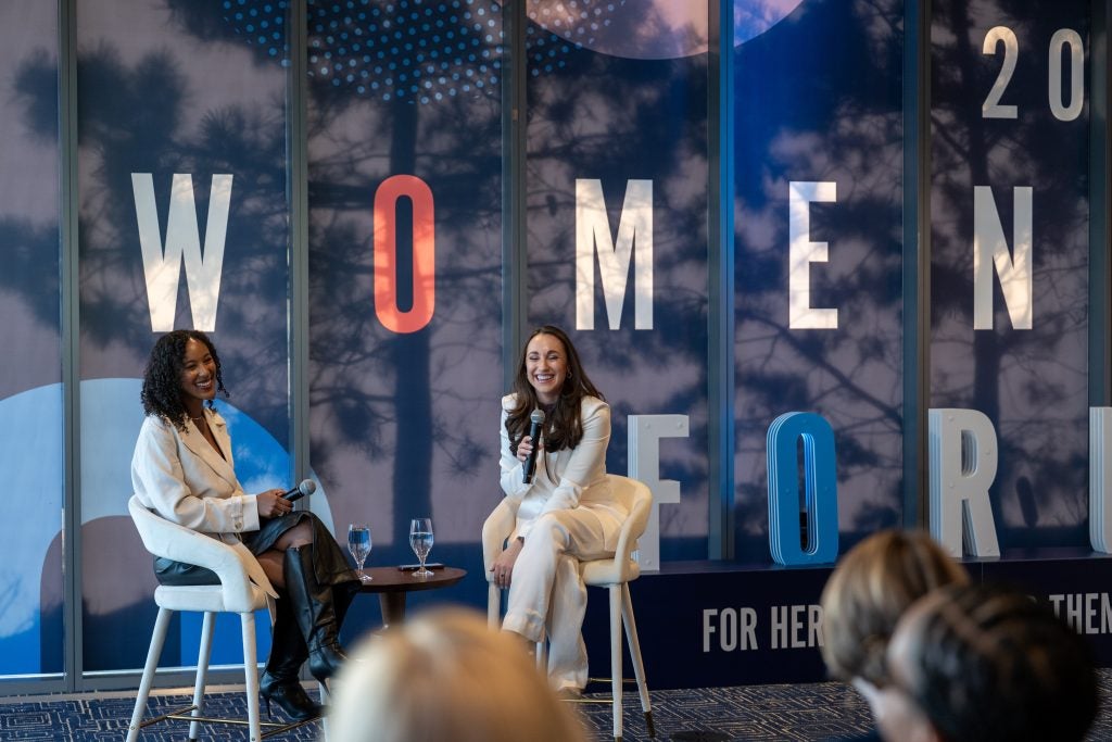 Two women sit in front of an audience, one is speaking into a microphone.