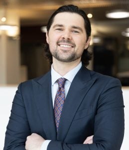 Headshot of Aubrey, wearing a navy suit, blue and orange patterned tie, and smiling.