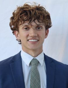 Headshot of Eli, wearing a navy suit and green plaid tie, smiling