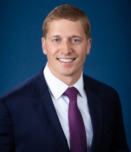 Headshot of Matt Yeager, wearing a navy suit, dark red polka dot tie, smiling