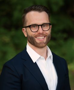 Headshot of Ken, wearing a black jacket, white shirt, glasses, and smiling