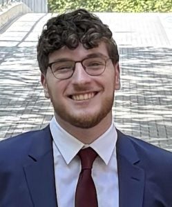 Headshot of Noah, wearing a navy suit and red tie, smiling