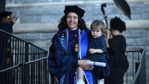 A mother in regalia holds her baby and diploma.
