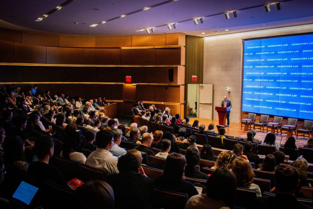 Rows of attendees in an auditorium listen to an onstage speaker