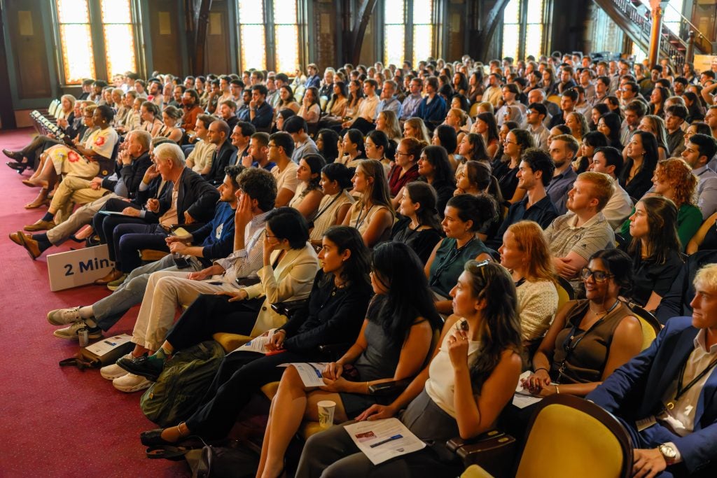 Rows of seated students fill a red-carpeted room with a wall of windows on one side.
