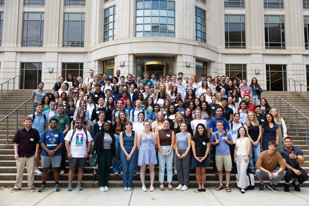 More than 100 students stand and smile on the steps of a school building.