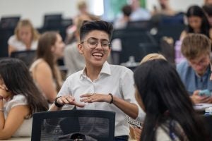A student with short dark hair and glasses smiles and gestures while talking to a classmate.