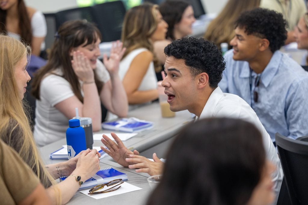 A student with dark hair speaks animatedly to a student with blonde hair. Spiral-bound books are on the table in between them.
