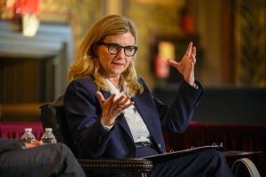A seated woman with light hair and glasses in a navy suit.