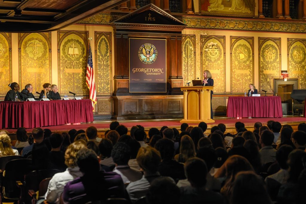 Six professors (five seated, one standing) onstage in a hall with ornate wall decor, while rows of students look on.