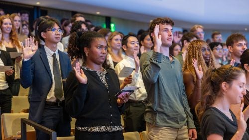Rows of standing students raise their right hands while reciting an oath