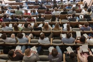 Students seated in rows are pictured from above. They are holding white paper handouts. 