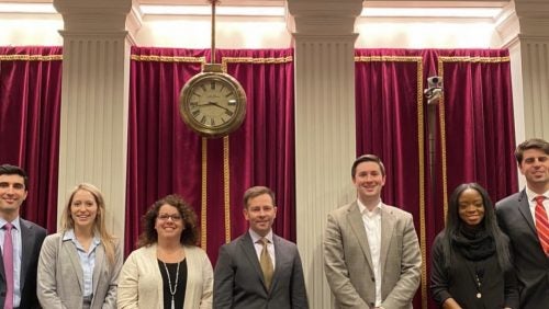 Seven people in suits smiling in front of columns and under a clock