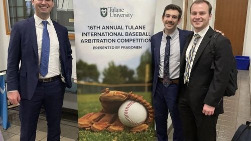 3 students in suits smiling in front of competition banner